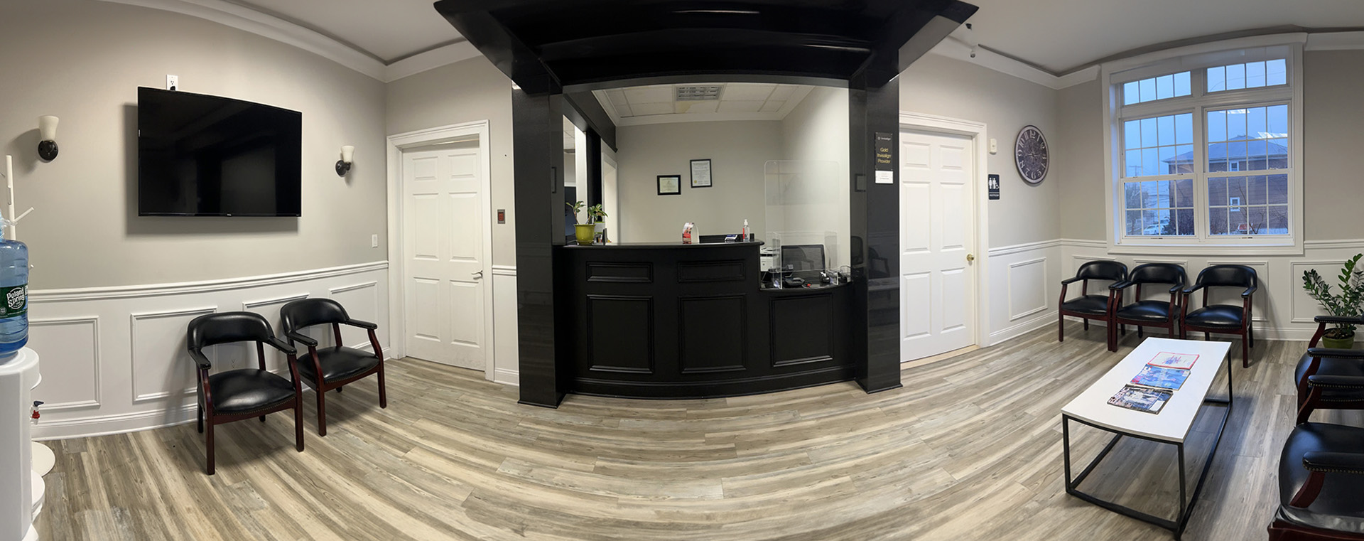 The image shows an interior space, possibly a waiting room or reception area within a building, with a tiled floor, a dark wood cabinet, a television screen mounted on the wall, a seating arrangement that includes chairs and a bench, and a large window allowing natural light to enter.