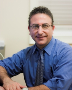 The image features a man with glasses, wearing a blue shirt and tie, sitting at a desk with a computer monitor.