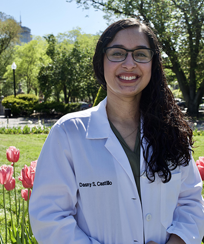 The image shows a person standing outdoors, wearing a white lab coat with text on it, dark glasses, and smiling at the camera. They have long hair and are wearing a name tag that reads  DONNY C. KELLY.  Behind them, there s a clear sky and some greenery.