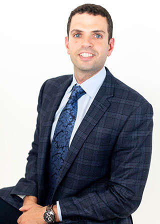 The image features a man wearing a suit and tie, posing with his hands clasped together, against a neutral background.