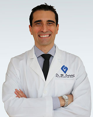 The image shows a man wearing a white lab coat with name tags on the left chest area, standing against a light background with his arms crossed and smiling at the camera.