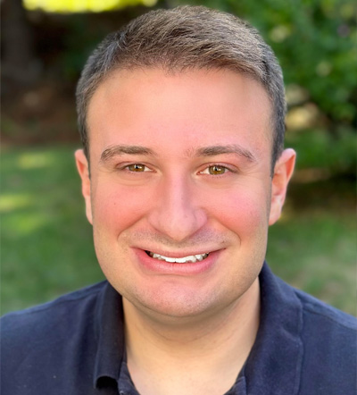 This image features a man with short hair smiling at the camera. He has a light skin tone and appears to be wearing a dark-colored shirt. The background suggests an outdoor setting, possibly a garden or park, with green foliage.