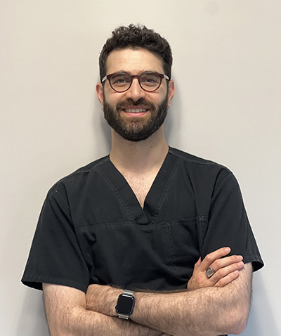 The image shows a man wearing glasses, standing against a wall with his arms crossed, dressed in a professional black shirt and dark pants, which suggests he might be a healthcare worker such as a doctor or nurse.