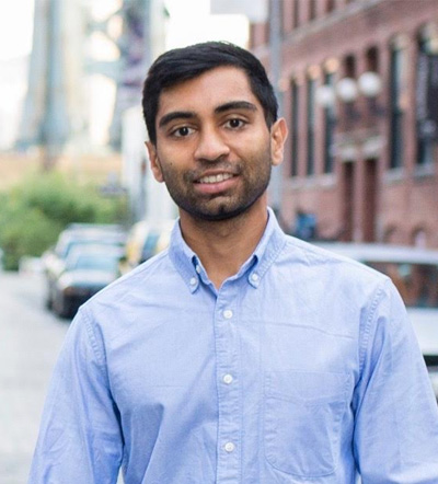 The image shows a man standing outdoors on a street with buildings behind him. He is smiling and wearing a light blue shirt, dark pants, and has short hair. There are no visible texts or logos in the image that provide additional context.