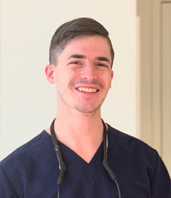 The image shows a smiling man wearing glasses, a blue shirt, and a necklace, standing indoors with white walls behind him. He has short hair and appears to be a professional, possibly a healthcare worker given his attire and the stethoscope visible around his neck.