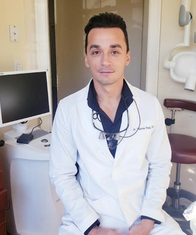 In the image, there is a man sitting in an office chair with his arms crossed, wearing a white lab coat, dark sunglasses, and a black shirt underneath, smiling slightly at the camera. He has short hair and appears to be professionally dressed, possibly for a dental or medical setting.