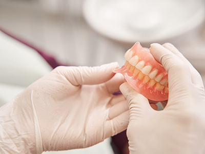 The image shows a dental professional wearing gloves and a mask, holding a pair of dentures and examining them closely.