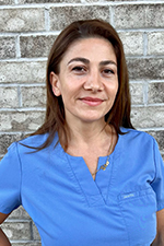 The image shows a woman wearing a blue scrub top, standing against a brick wall with her arms crossed. She appears to be outdoors during daylight.
