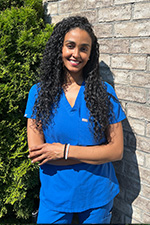The image features a woman wearing scrubs, standing against a brick wall with her arms crossed.