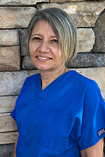 A woman wearing a blue scrub uniform stands against a stone wall background.