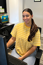 A woman sitting at a desk with a computer monitor, smiling towards the camera.