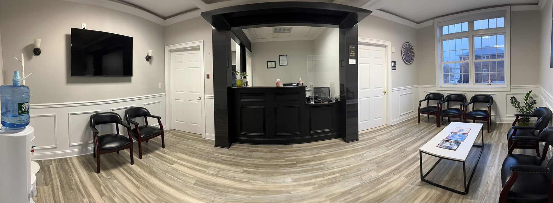 The image shows an interior view of a waiting room with a modern design featuring a black reception desk, a white wall, a wooden floor, and various chairs arranged around the room.