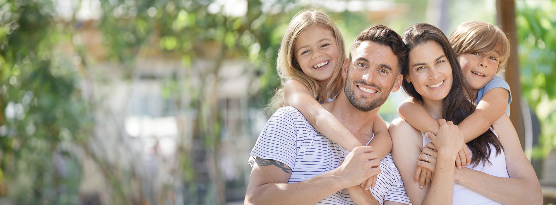 The image shows a family of four, including two adults and two children, posing for a photo outdoors with trees in the background.