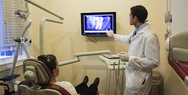 A dentist in a white lab coat conducting an examination on a patient seated in a dental chair, with a large monitor displaying an X-ray of their mouth.