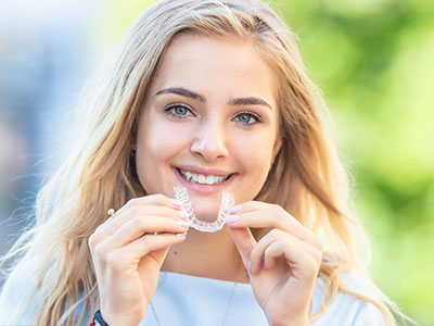 A young woman with blonde hair is smiling at the camera while holding up a clear dental retainer with her hands, showcasing its transparent material against a blurred outdoor background.