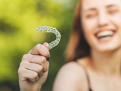 An adult female holds up a clear plastic dental retainer with her right hand while smiling at the camera.