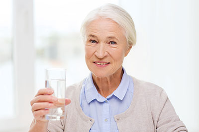 The image features an elderly woman holding a glass of water with both hands, smiling at the camera, wearing glasses, a light blue shirt, and dark pants.