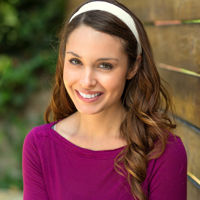 The image shows a woman with long brown hair smiling at the camera while posing for a portrait with a wooden fence in the background.