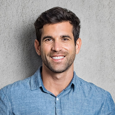 A young man with short hair, wearing a blue denim shirt, is smiling and posing against a light grey background.