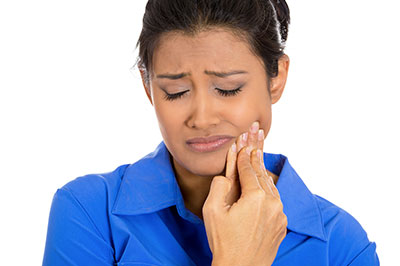 A woman with dark skin and hair, wearing a blue blouse, is looking down at her hand while holding something small between her fingers, with an expression of concern or worry on her face.