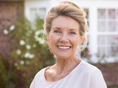 A smiling older woman with short blonde hair stands outdoors, wearing a white top, against a backdrop of a house and greenery.