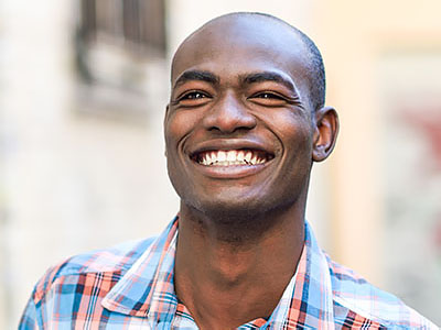 The image shows a man smiling broadly at the camera, with his mouth open, revealing his teeth. He appears to be outdoors, possibly in an urban setting, given the architectural elements visible behind him.