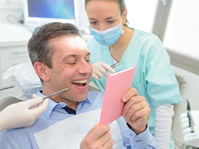 A man sitting in a dental chair holding a pink card, with a woman standing behind him, smiling at the camera, and another person looking on.