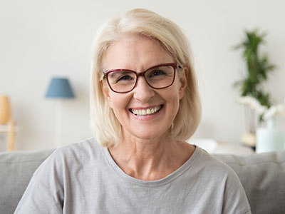 The image features a woman with short blonde hair, wearing glasses and a light-colored top, smiling and looking directly at the camera. She has a neutral background that includes a lamp and a partial view of a white wall.