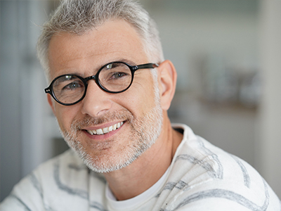 The image shows a smiling middle-aged man with short gray hair, wearing glasses and a white shirt, looking directly at the camera.