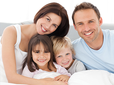 The image shows a family of four people lying together on a bed with smiles on their faces.