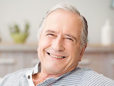 The image shows a smiling older man with gray hair, wearing a blue shirt, seated comfortably in a chair, looking directly at the camera.