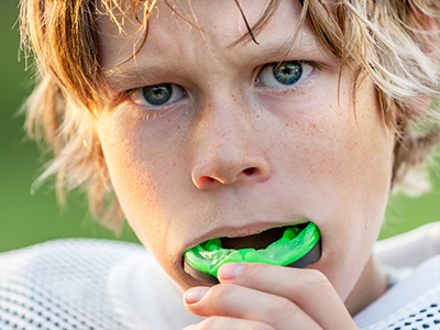 A young boy with blonde hair and blue eyes, wearing a football jersey, holding a toothbrush with green bristles in his mouth.