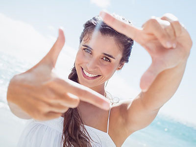 The image shows a woman with long hair smiling at the camera while holding up her index finger against the sky.